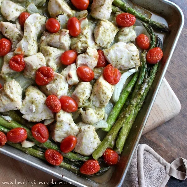 Angled photo of Lemon Pesto Sheet Pan Chicken with Asparagus and Tomatoes sitting on a cutting board with a pot holder