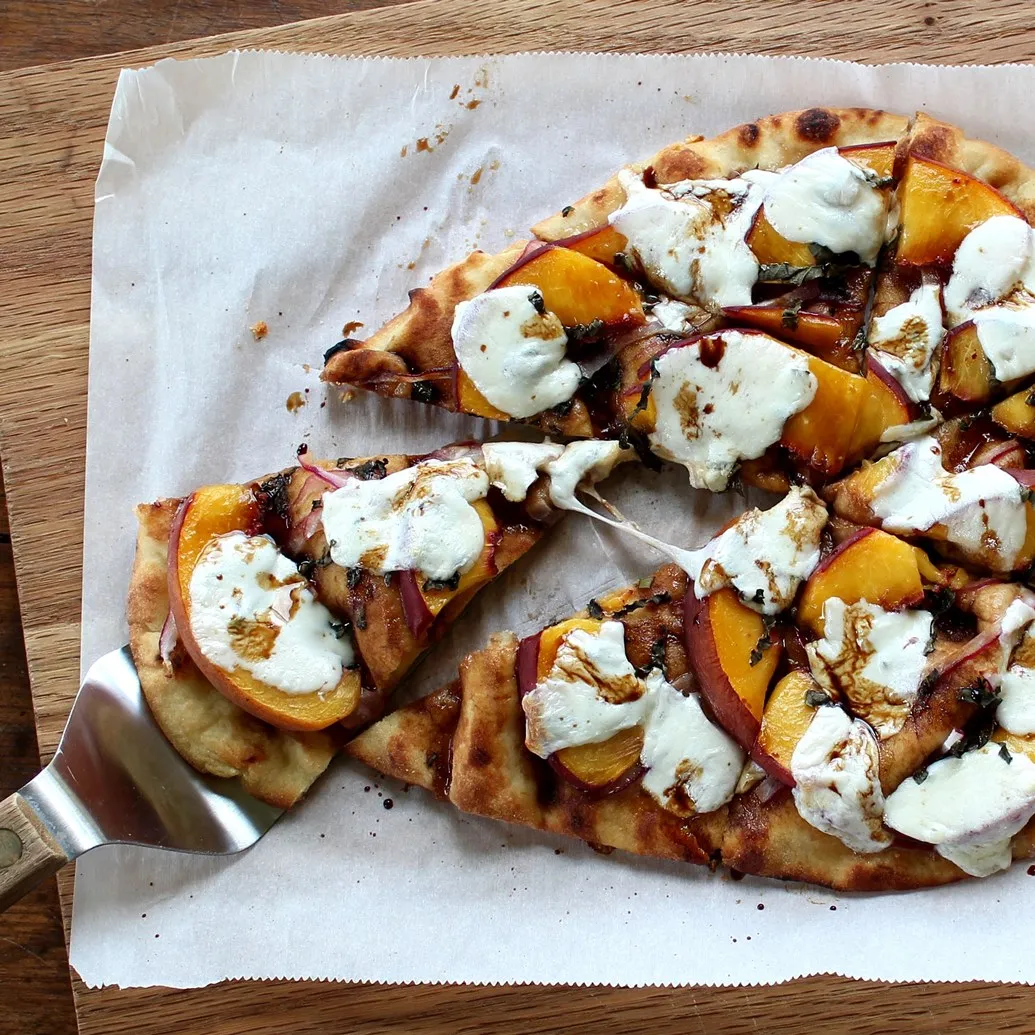 a pie server holding up a piece of Naan Pizza with Peaches, Basil, and Fresh Mozzarella