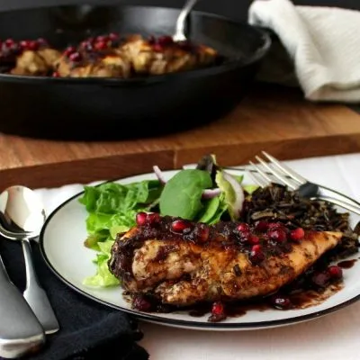 Pomegranate-Cranberry Chicken on a white and black plate with a black napkin and utensils