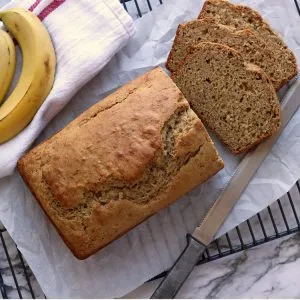 Banana bread with three slices cut off, sitting on a cooling rack with a knife