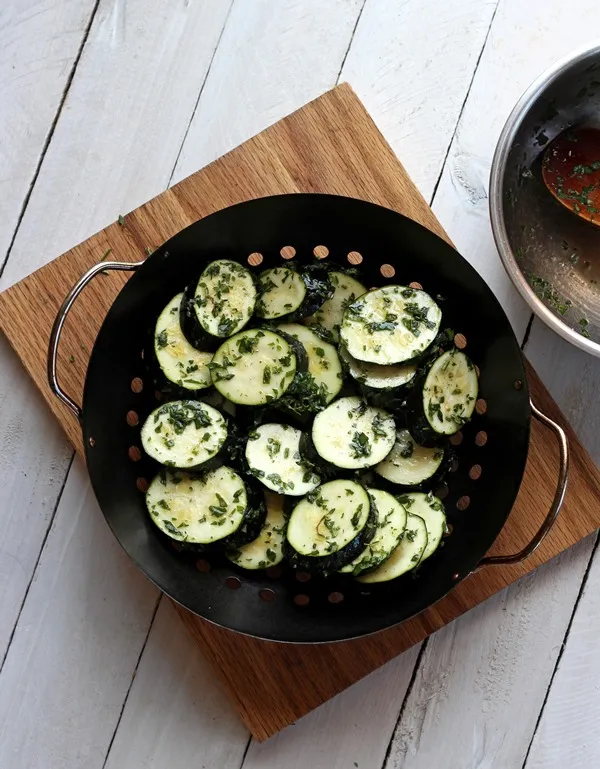 Zucchini sliced and chopped fresh herbs in a grill basket on a wood cutting board