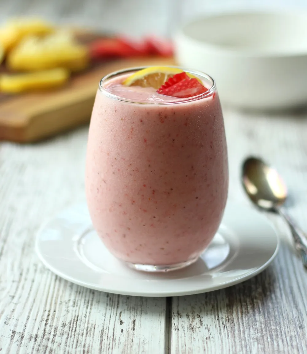 pink smoothie in clear glass with lemons and strawberries on cutting board in background