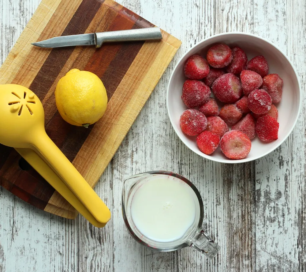 cup of milk, frozen strawberries in a white bowl, and lemon with a knife and juicer on cutting board