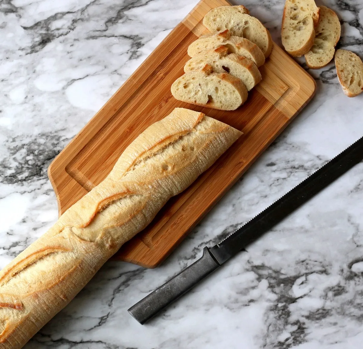 Partially cut baguette on wood cutting board with serrated knife nearby
